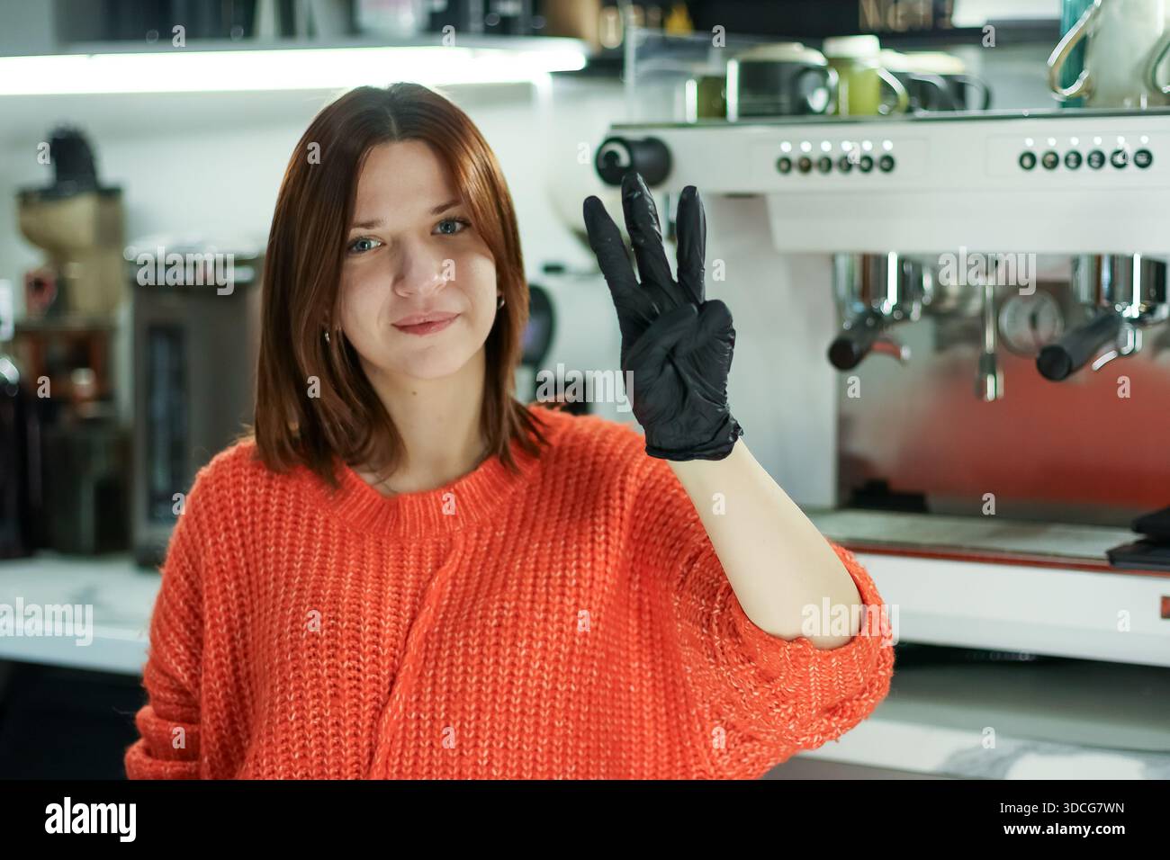 Femme souriante en pull tricoté et gants noirs gestes trois doigts derrière la machine à café dans l'intérieur moderne du café. Banque D'Images