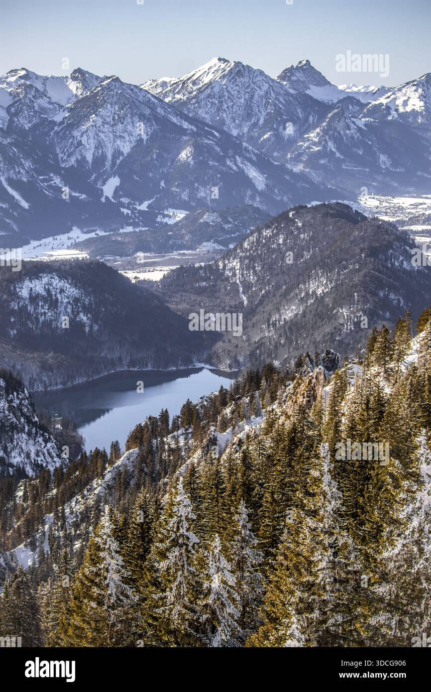 Vue sur des sapins poussiéreux de neige accrochés à des pentes rocheuses, un lac serein reflétant le ciel, et des montagnes majestueuses et enneigées transpercant l'horizon, BAV Banque D'Images