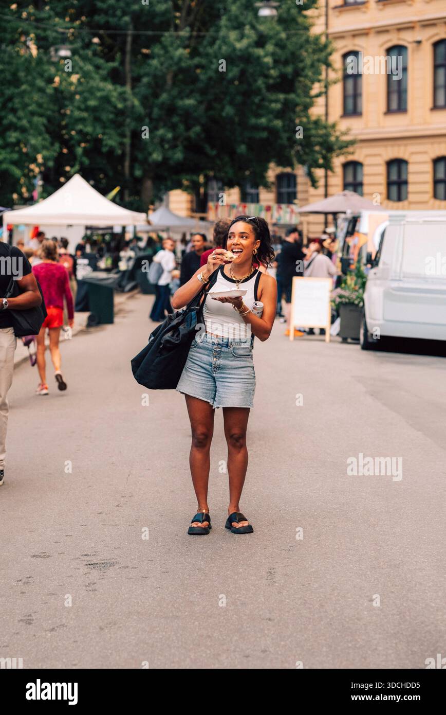 Pleine longueur de femme mangeant apéritif tout en se tenant sur la route à la fête de rue Banque D'Images