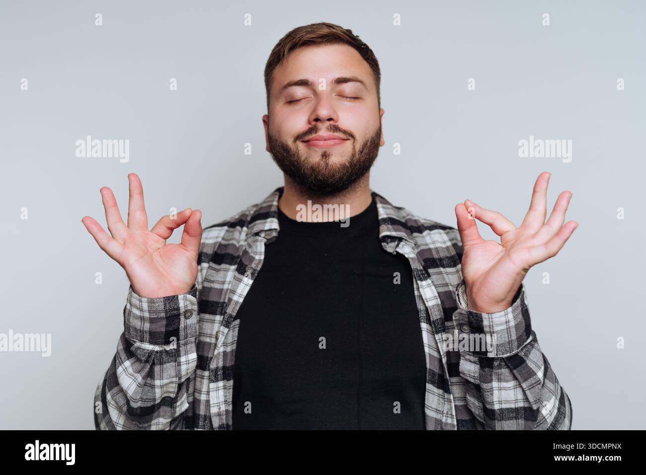 Un homme est assis dans une pièce calme, les yeux fermés, pratiquant la méditation. Il tient ses mains dans un geste de paix tout en portant une chemise à carreaux et un Banque D'Images