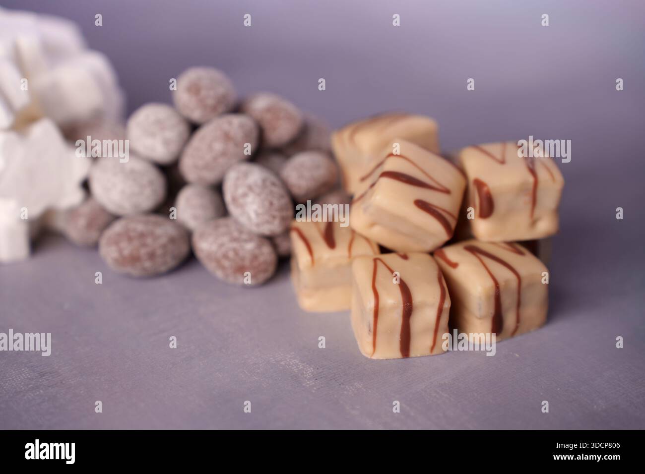 gros plan de confiseries assorties avec des cubes de caramel au filet de chocolat et des amandes enrobées de sucre sur une surface douce de lavande avec un éclairage doux, Banque D'Images