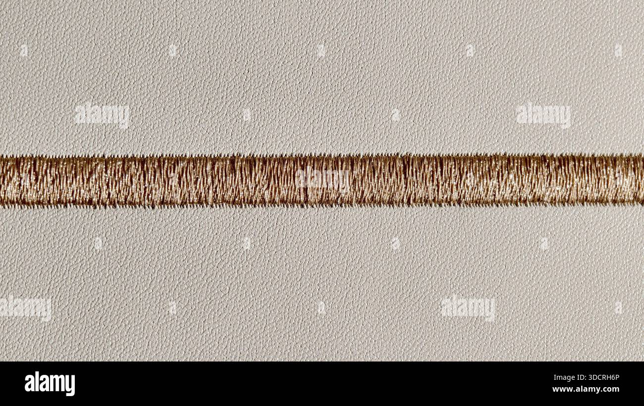 Gros plan d'un point de broderie doré horizontal sur une surface en cuir crème texturé. Le détail fil fin et le grain cuir créent un style sophistiqué et haut Banque D'Images