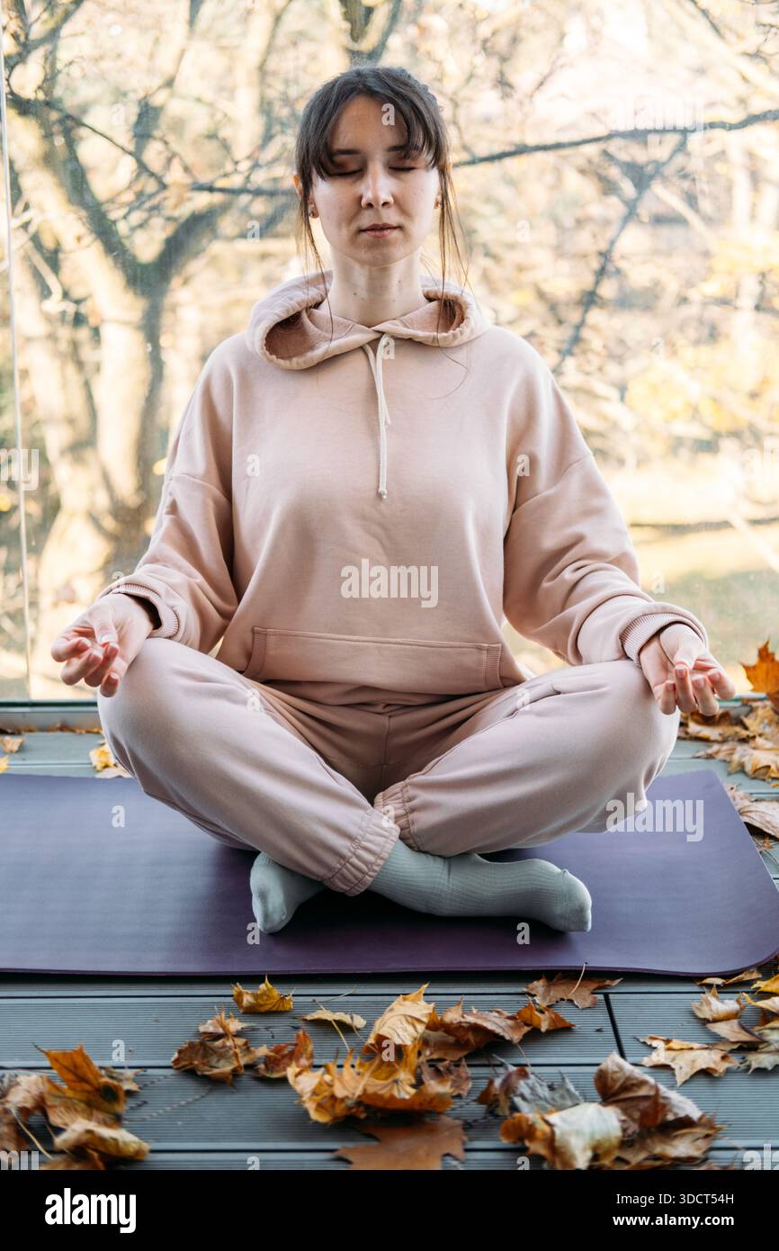 La femme est assise les yeux fermés dans la pose méditative sur le balcon pendant l'automne. Habitudes de soins personnels, sanctuaire de bien-être à domicile, style de vie tranquille. Banque D'Images
