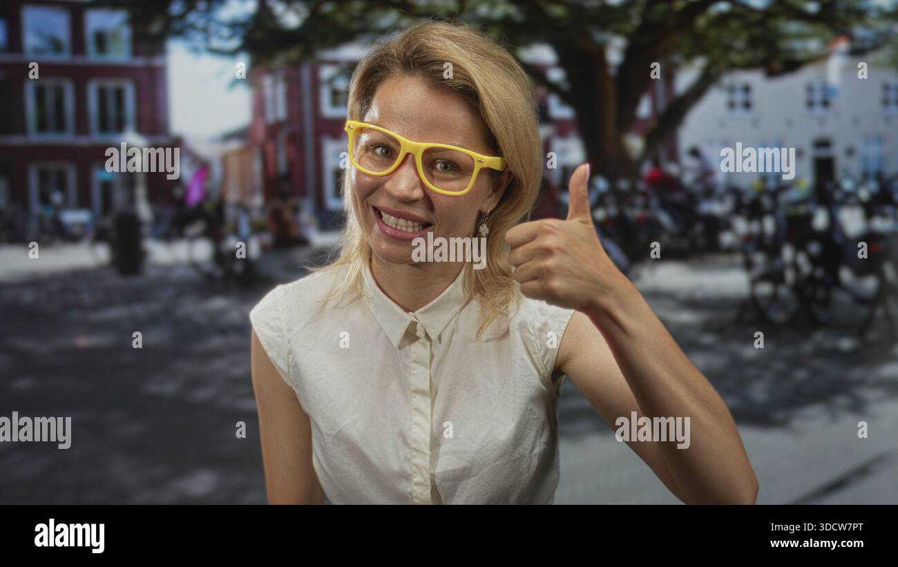 Jeune femme blonde souriante donne un geste du pouce vers le haut sur une rue urbaine animée en plein jour pendant la journée ensoleillée ; optimisme. Banque D'Images