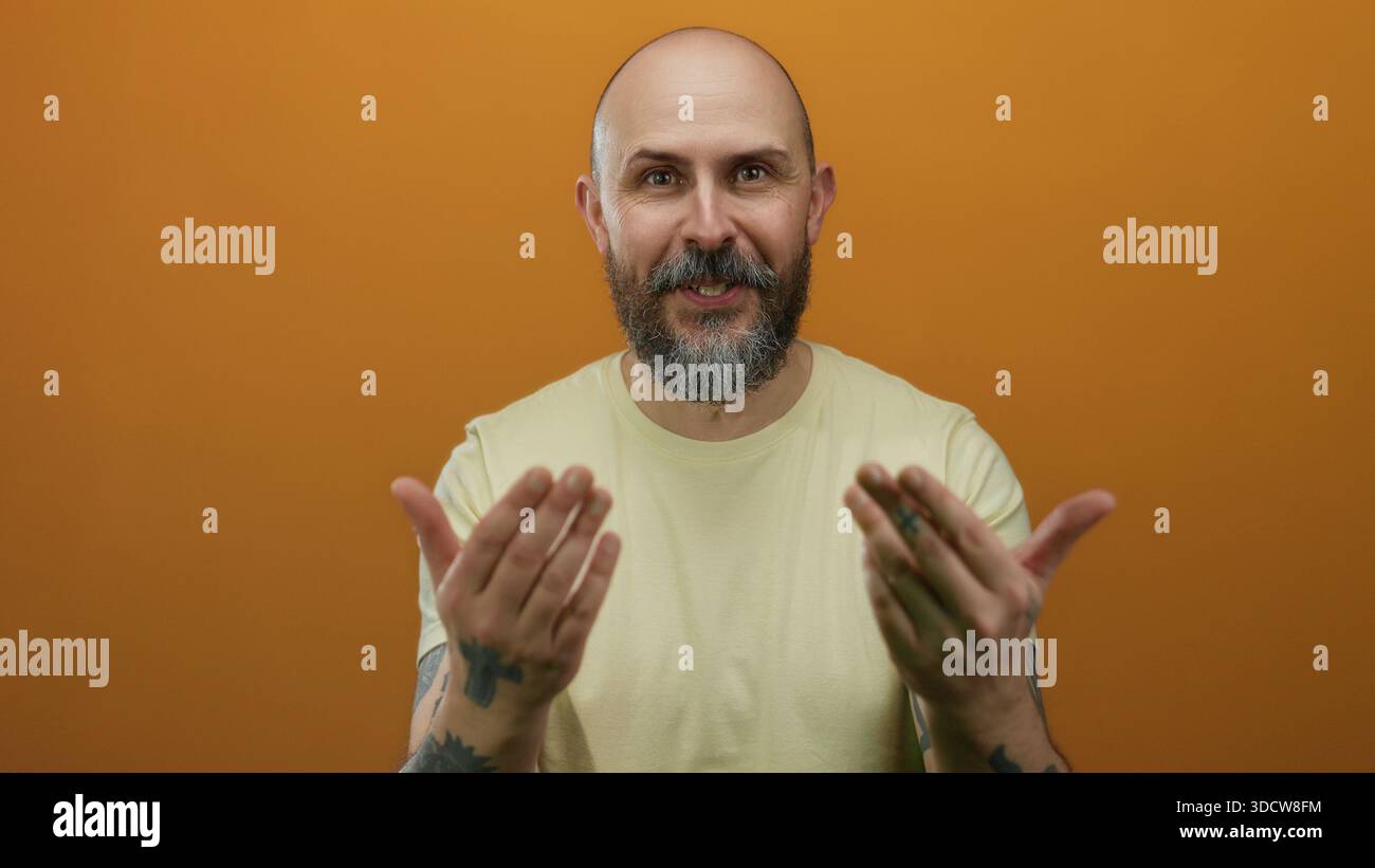 Homme barbu chauve gestes avec les mains contre le mur orange mettant en vedette des tatouages et l'expression invitante, portant une chemise jaune dans un set de studio isolé Banque D'Images