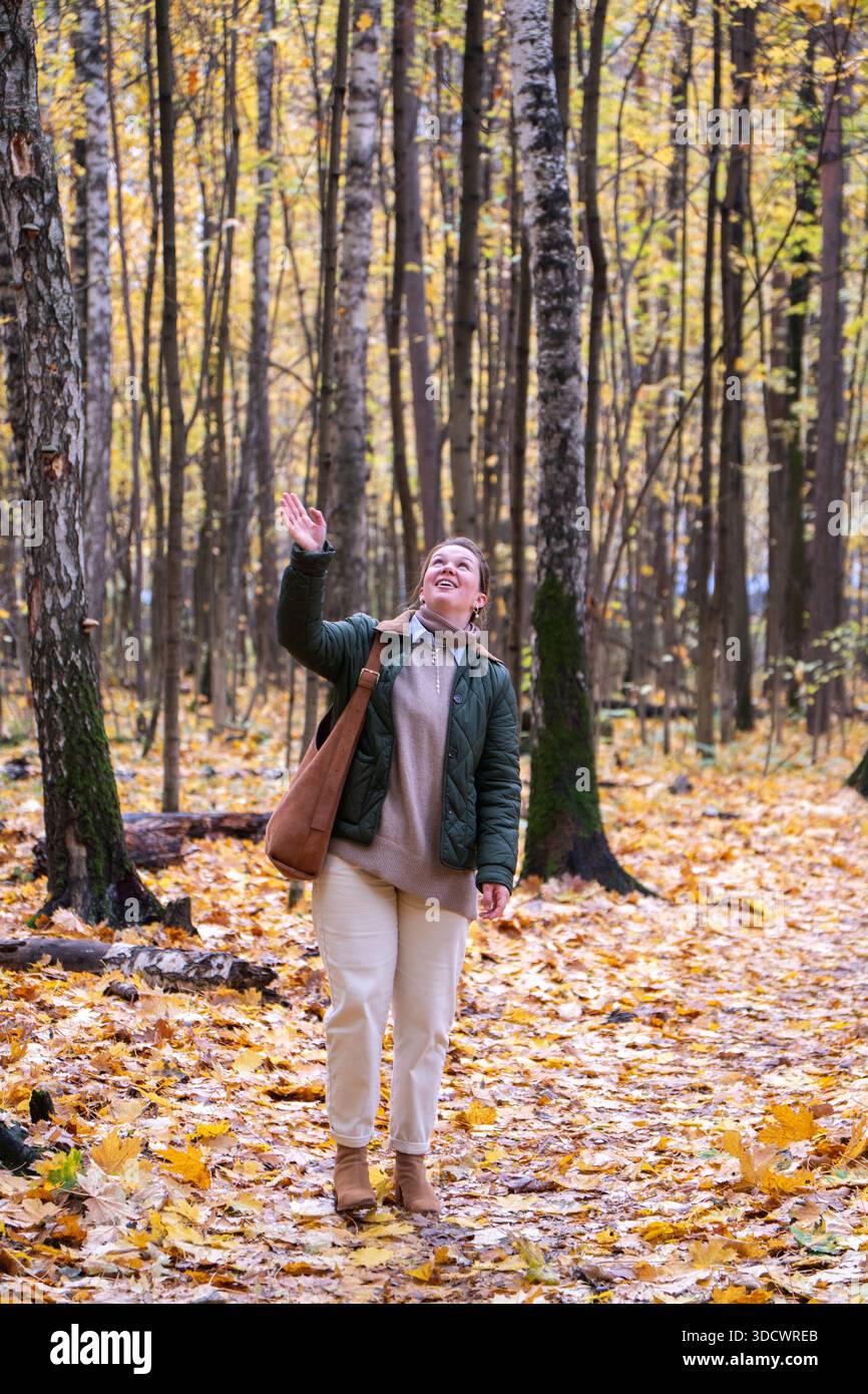 Femme souriante marche à travers une forêt d'automne, feuilles tombées jaunes sous les pieds, grands arbres, couleurs chaudes, humeur joyeuse et détendue Banque D'Images