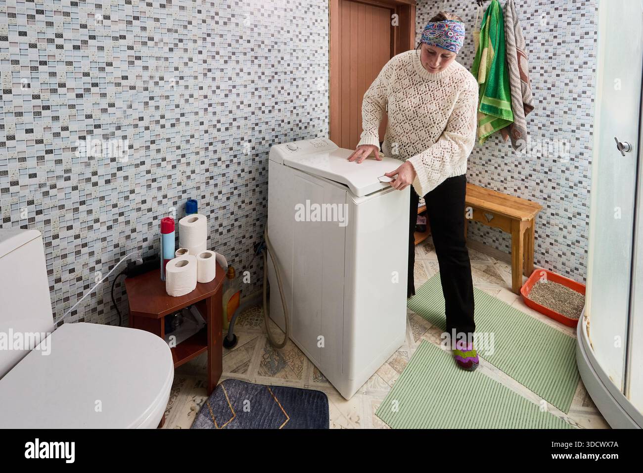 Femme au foyer ferme le couvercle de la machine à laver automatique à chargement par le haut installée dans la salle de bain entre les toilettes et la cabine de douche. Banque D'Images