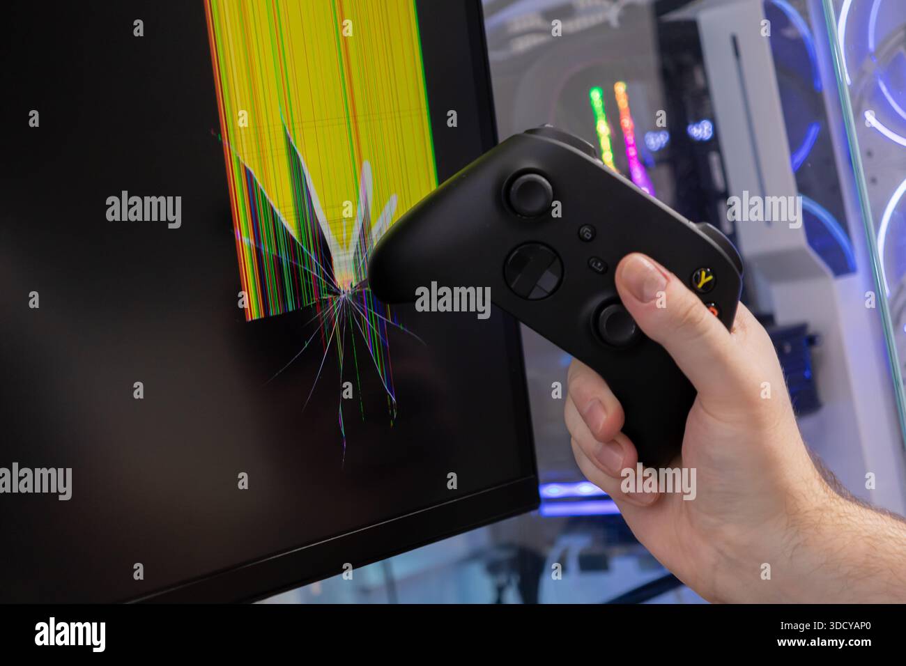 Gros plan d'une main tenant une manette de jeu impactant un écran d'ordinateur endommagé, affichant un verre fracturé et des lignes de pixels éclatantes, illustrant p. Banque D'Images