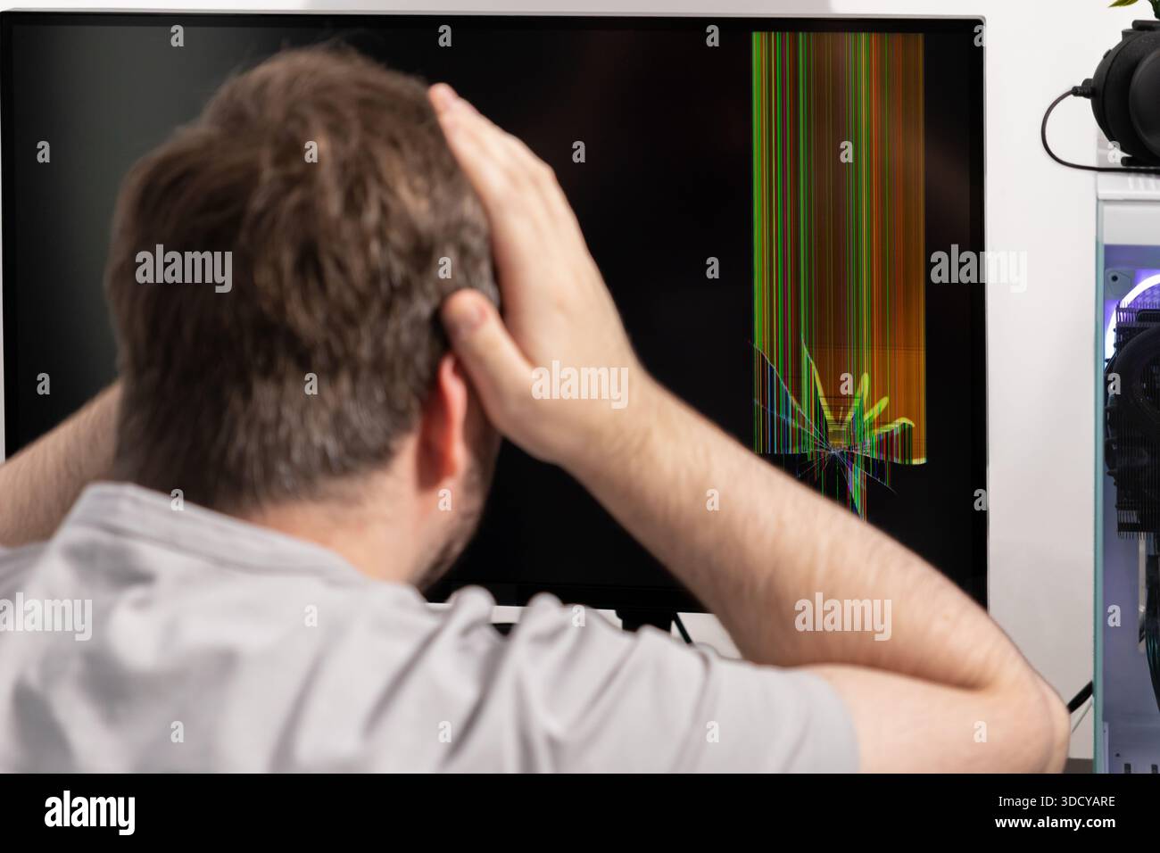 L'homme détraumatisé observe un moniteur d'ordinateur de bureau endommagé avec des lignes verticales colorées et une fissure d'écran visible, mettant en évidence une défaillance matérielle inattendue Banque D'Images