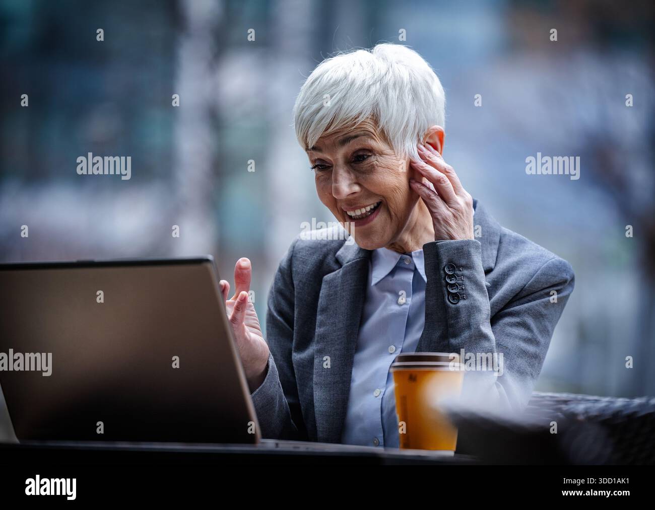 Femme d'affaires s'assoit à une table dans un café extérieur et ayant un appel vidéo sur ordinateur portable. Elle est habillée d'un costume et a les cheveux courts. Banque D'Images