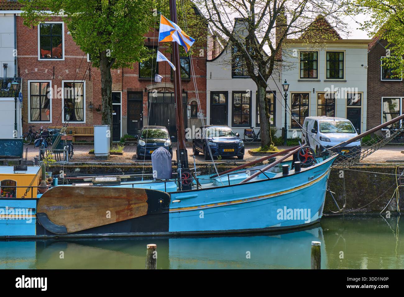 Une barge bleue du type Klipper avec un grand leeboard se trouve sur un canal à Den Briel pays-Bas avec des drapeaux hollandais agitant dans le fond les voitures sont p Banque D'Images
