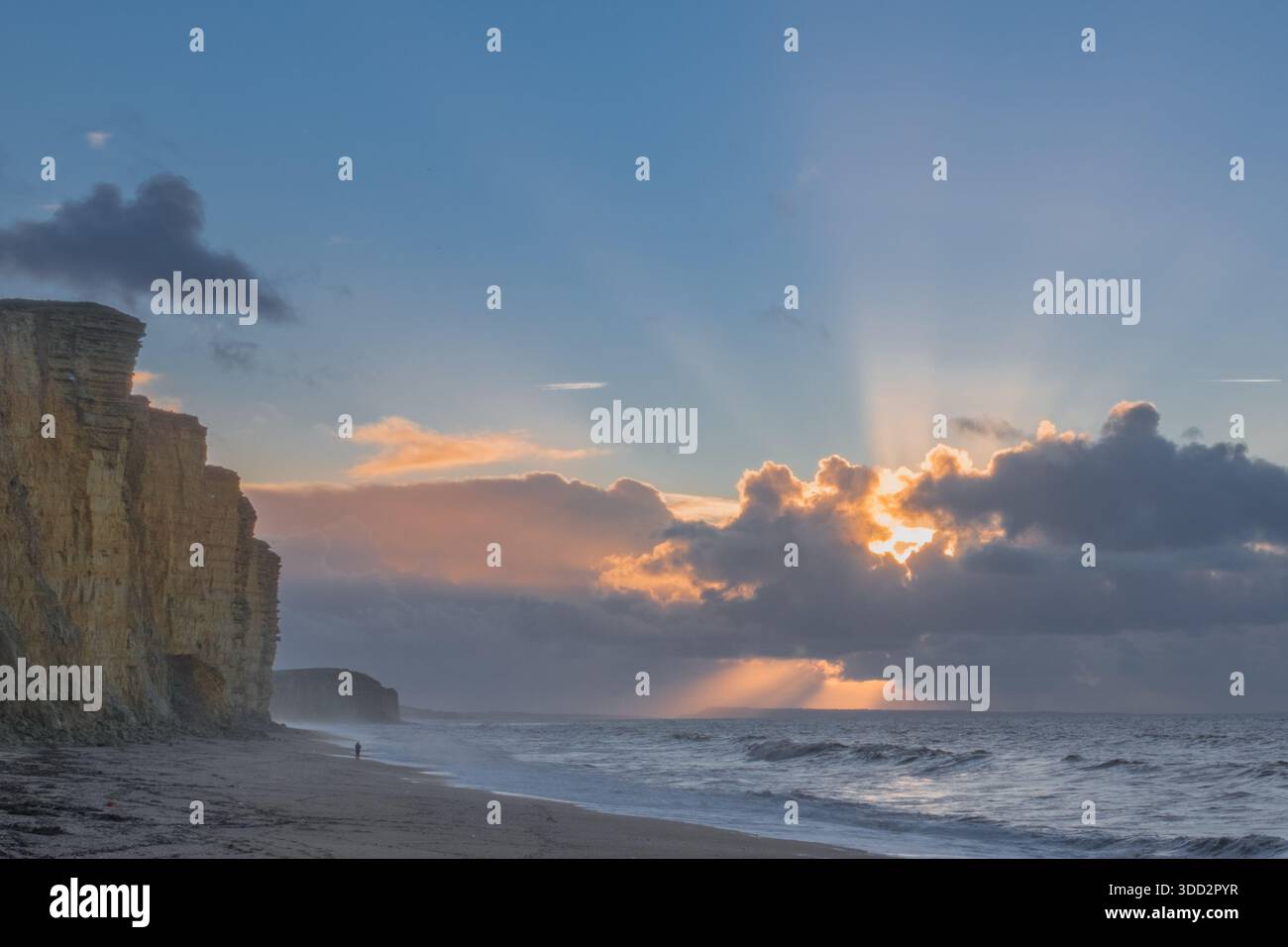 Les spectaculaires falaises de West Bay, éclairées par le lever du soleil avec un marcheur solitaire marchant le long de la plage sous des falaises très instables. Banque D'Images