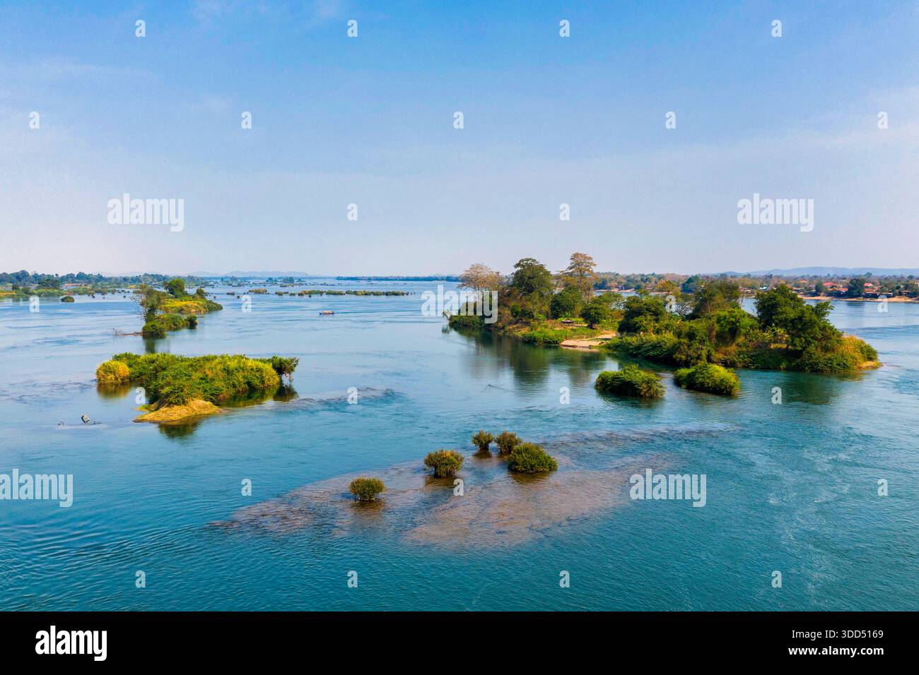 Un large tronçon du fleuve Mékong coule devant des îlots verts éparpillés et une rive bordée d'arbres dans la région des 4000 îles du sud du Laos. La lumière du jour claire et l'eau calme créent un paysage riverain spacieux avec un feuillage éclatant et des reflets doux. Banque D'Images