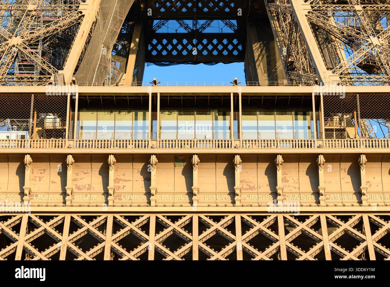 Vue directe sur le premier étage de la tour Eiffel, avec façade en fer vieilli, noms de scientifiques gravés et pavillon moderne en verre baigné de lumière dorée. Banque D'Images