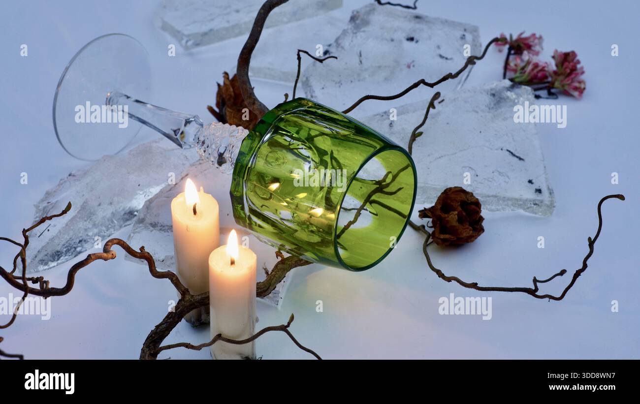 Un verre de cristal tombé entre des brindilles, des fleurs fanées et des morceaux de glace. Deux bougies. Banque D'Images