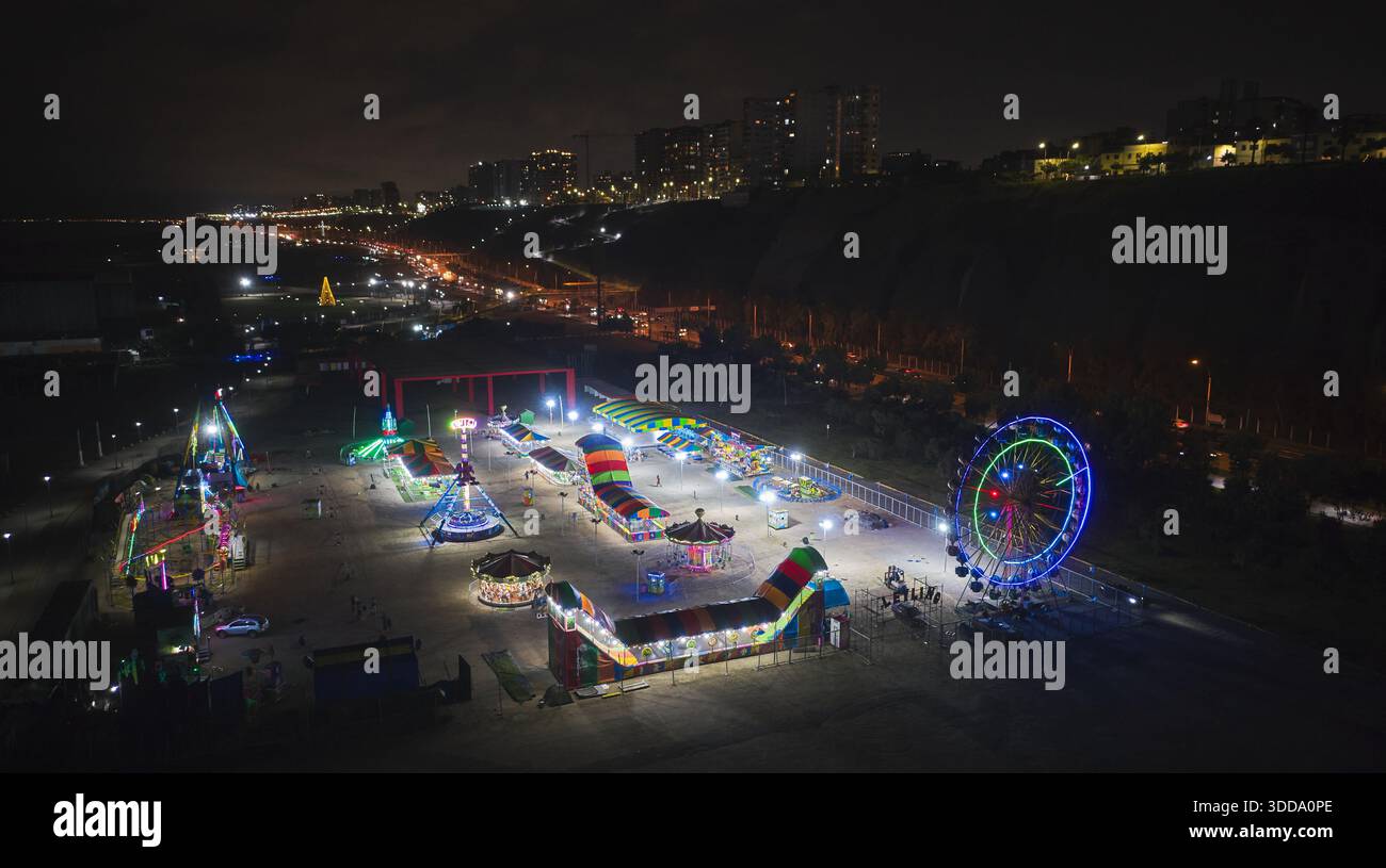 Parc d'attractions lumineux avec une grande roue et des manèges colorés illuminant la nuit au bord de la mer Banque D'Images