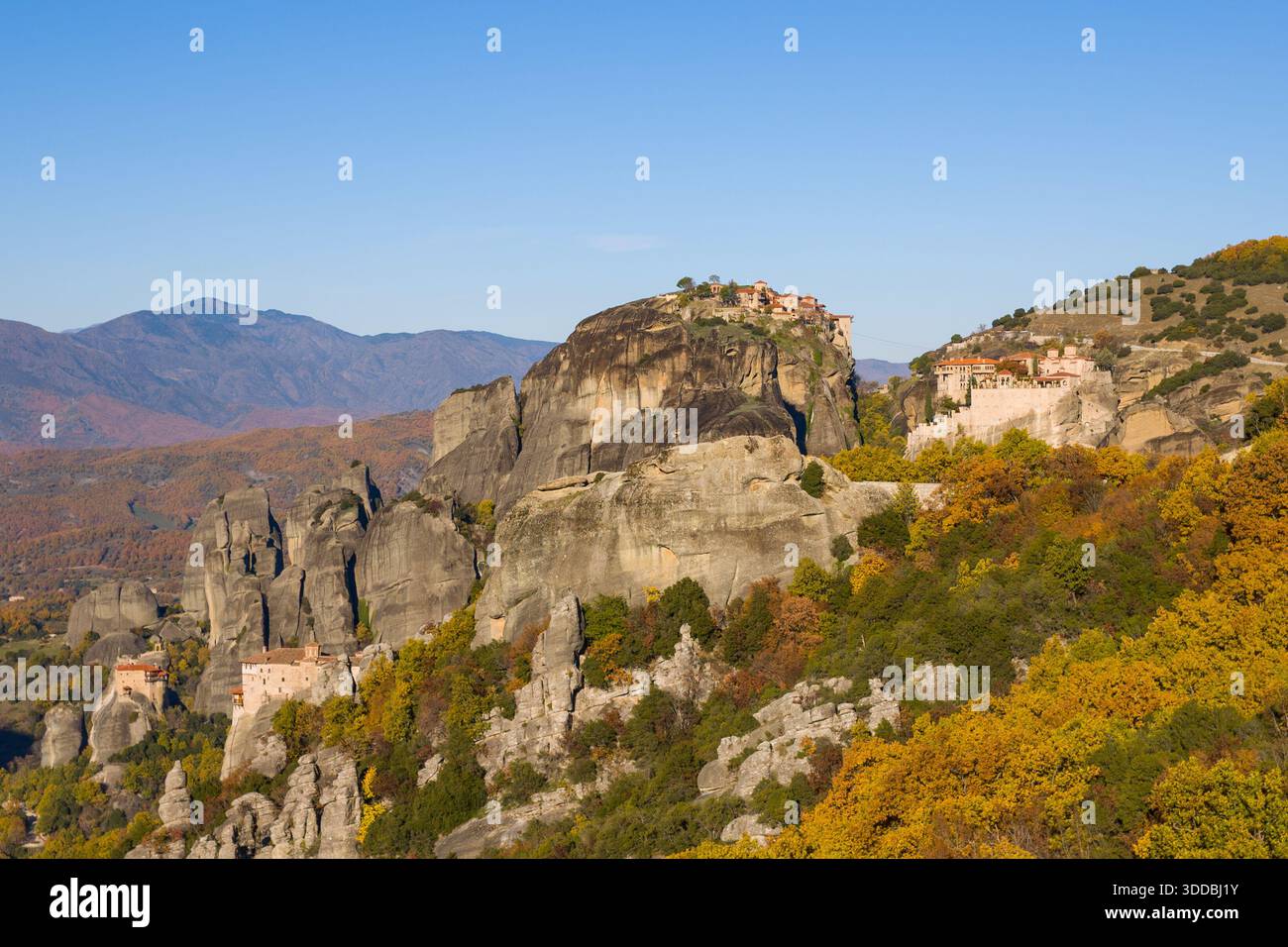 Vue panoramique sur les Météores en Grèce, avec d'anciens monastères perchés sur d'imposantes falaises de grès entourées de feuillage d'automne et de montagnes lointaines sous un ciel bleu clair. Le paysage met en valeur des formes géologiques saisissantes et des couleurs saisonnières vibrantes. Banque D'Images