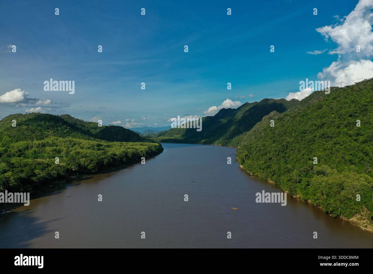 Un large tronçon du fleuve Mékong coule entre des collines verdoyantes densément boisées sous un ciel bleu vif avec des nuages éparpillés. La scène présente une végétation tropicale riche, des pentes douces et une eau tranquille en Asie du Sud-est. Banque D'Images