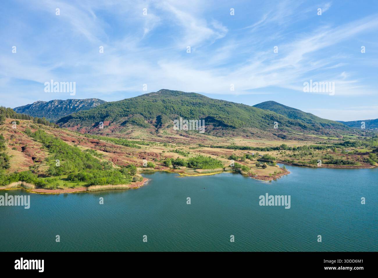 Vue panoramique sur le lac de Salagou avec des collines verdoyantes s'élevant au-dessus de la terre rouge et de l'eau bleue calme, situé sous un ciel lumineux et légèrement nuageux. Le paysage présente des couleurs naturelles vibrantes et une atmosphère paisible et ouverte. Banque D'Images
