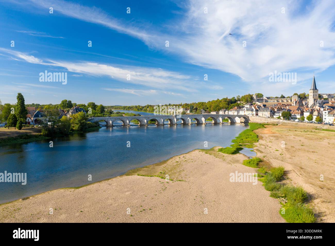 Vue imprenable sur la Charite-sur-Loire dans le centre de la France, avec un pont de pierre historique traversant la Loire, une large rive de sable au premier plan et la flèche de l'église de la ville s'élevant au-dessus des toits traditionnels. Des nuages tortueux s'étendent à travers un ciel bleu vif, mettant en valeur le paysage tranquille de la rivière. Banque D'Images