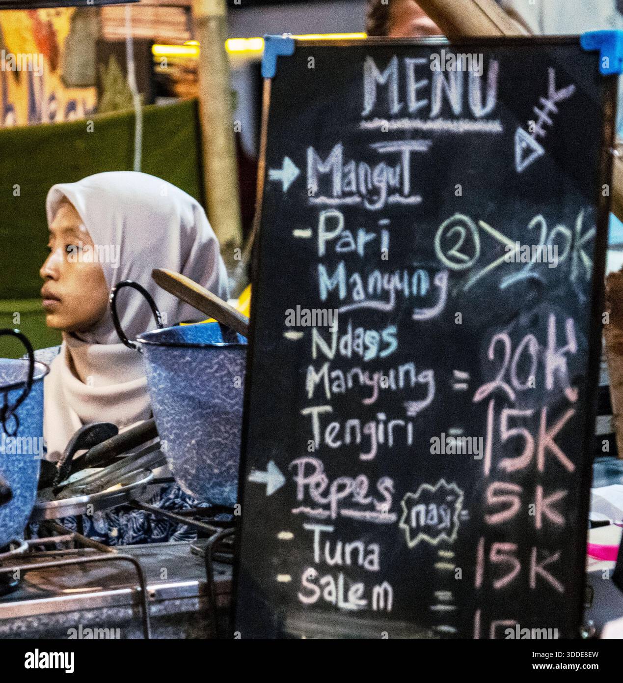 Une femme en hijab se tient près d'un stand de nourriture avec un menu au tableau noir. Banque D'Images