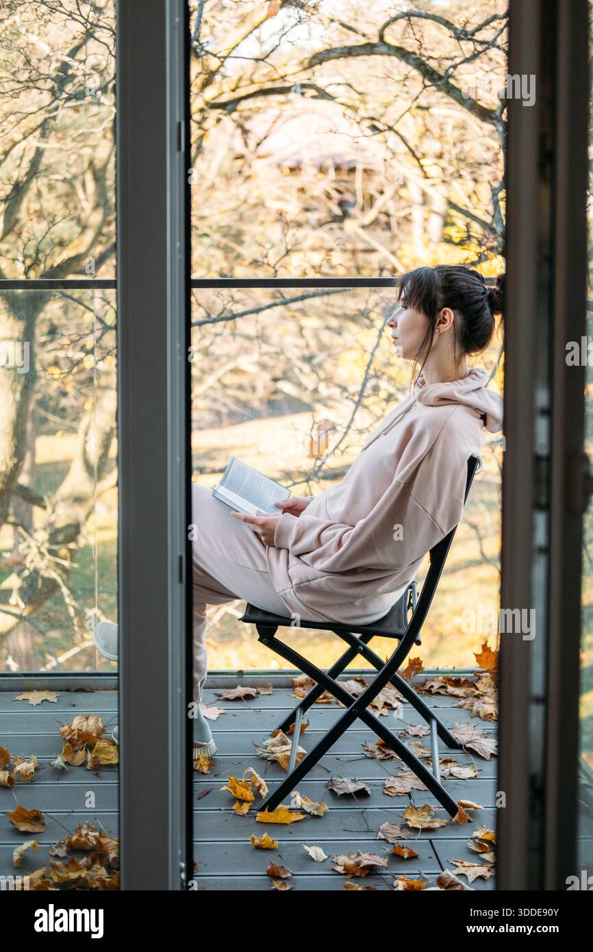 Femme assise sur la chaise sur le balcon lisant livre entouré de feuilles d'automne tombées. Vie lente, solitude urbaine, moments conscients, plaisirs simples. Banque D'Images