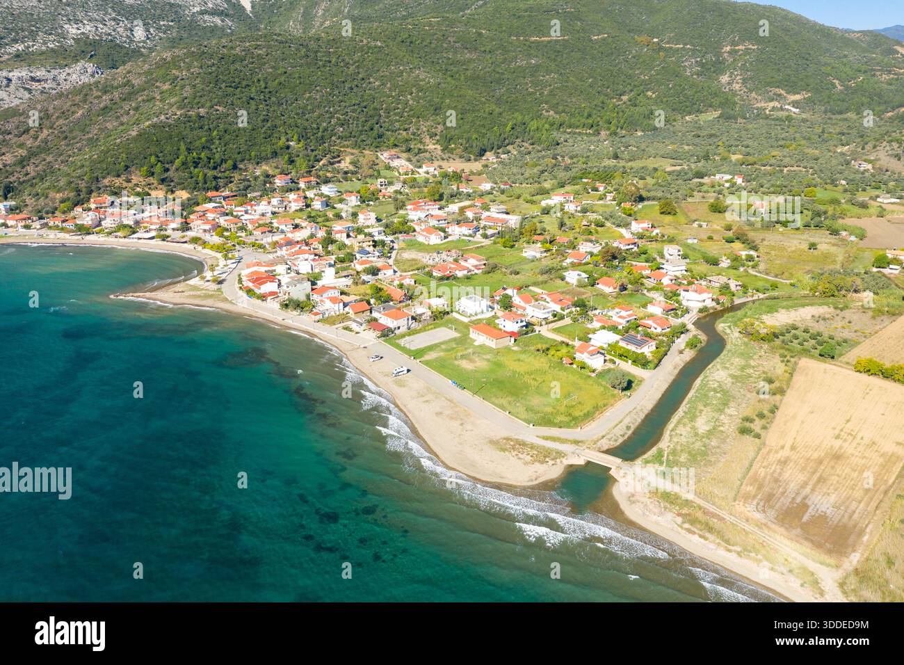 Une vue en grand angle de Kato Vasiliki montre un groupe de maisons au toit rouge le long d'un rivage incurvé, bordé par des eaux turquoises et des collines verdoyantes sous la lumière du soleil. Le paysage comprend un petit canal, des champs ouverts et une atmosphère méditerranéenne tranquille. Banque D'Images