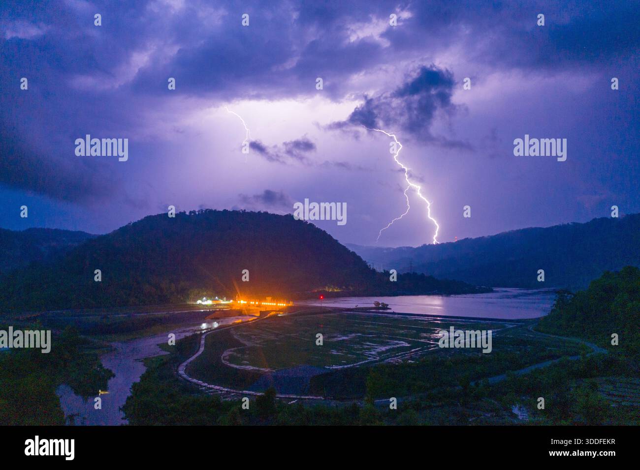 Un éclair éclatant illumine les nuages orageux spectaculaires au-dessus d'un réservoir isolé et de champs en terrasses, avec des lumières incandescentes près du rivage qui se reflètent sur l'eau. La scène capture l'énergie dynamique et l'atmosphère sombre d'un orage nocturne dans un paysage montagneux. Banque D'Images
