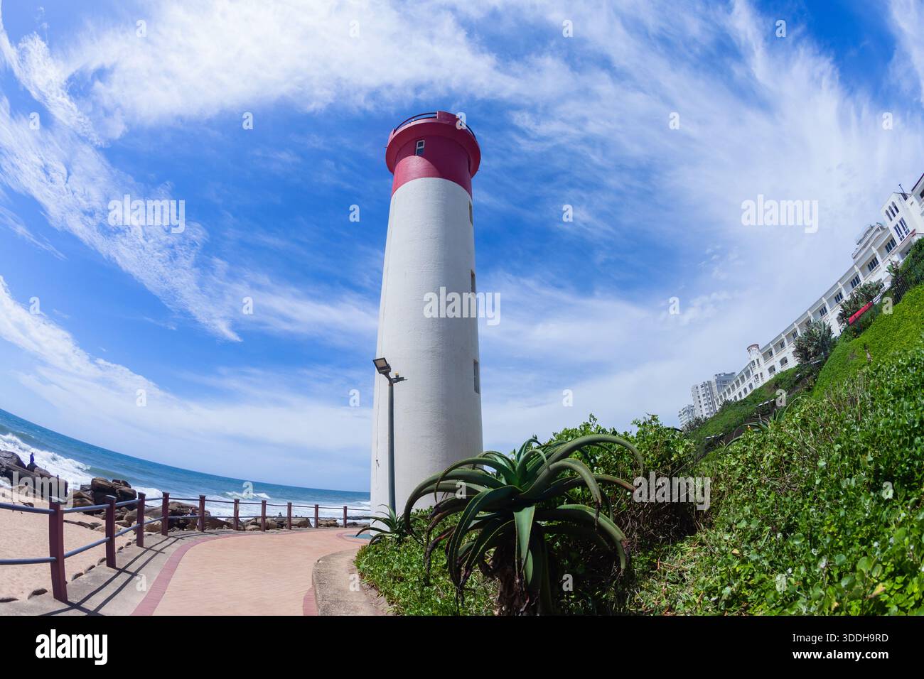 Phare le long de la côte de plage rocheuse se dresse haut avec de la peinture rouge et blanche à côté des appartements de construction et le ciel bleu d'été. Banque D'Images