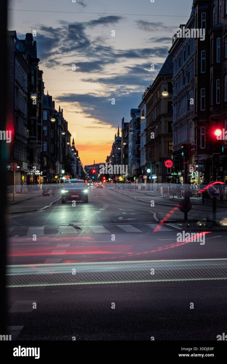 Longue exposition de la circulation sur un boulevard de la ville au crépuscule, avec des traînées lumineuses, des lampadaires et des bâtiments historiques sous un ciel de coucher de soleil coloré. Banque D'Images