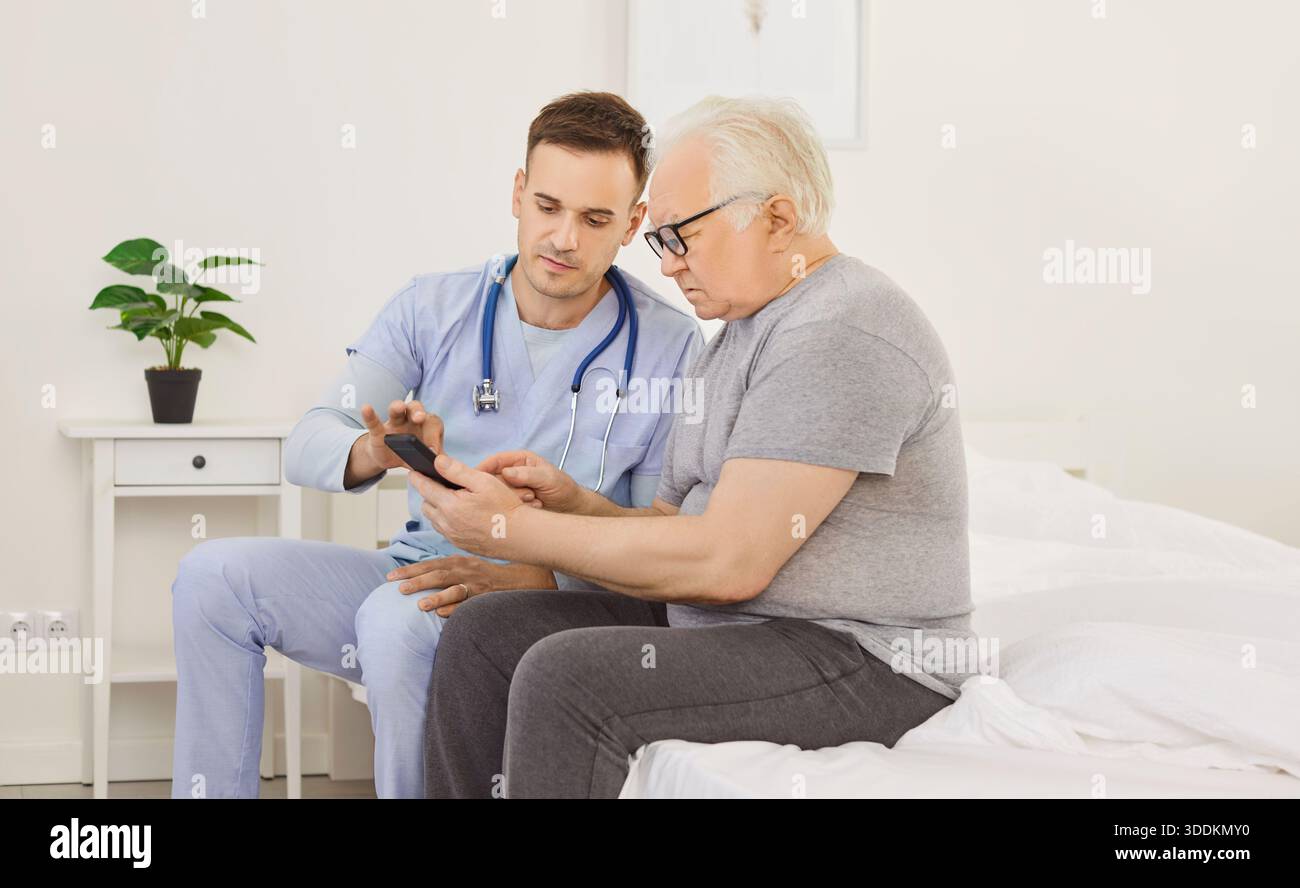 Jeune soignant masculin enseignant à une personne âgée à manipuler avec un téléphone portable assis sur le lit dans la clinique. Banque D'Images