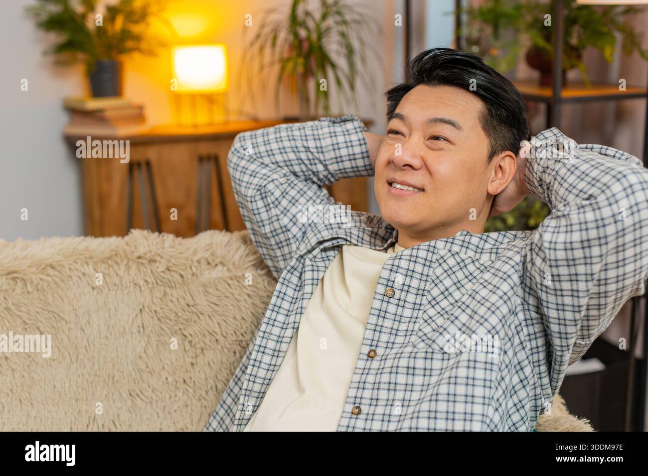Heureux homme chinois d'âge moyen insouciant assis confortablement avec la main derrière la tête sur le canapé dans le salon à la maison. Souriant mec coréen se relaxant sur le canapé pensant regarder autour dans la chambre de l'appartement. Banque D'Images