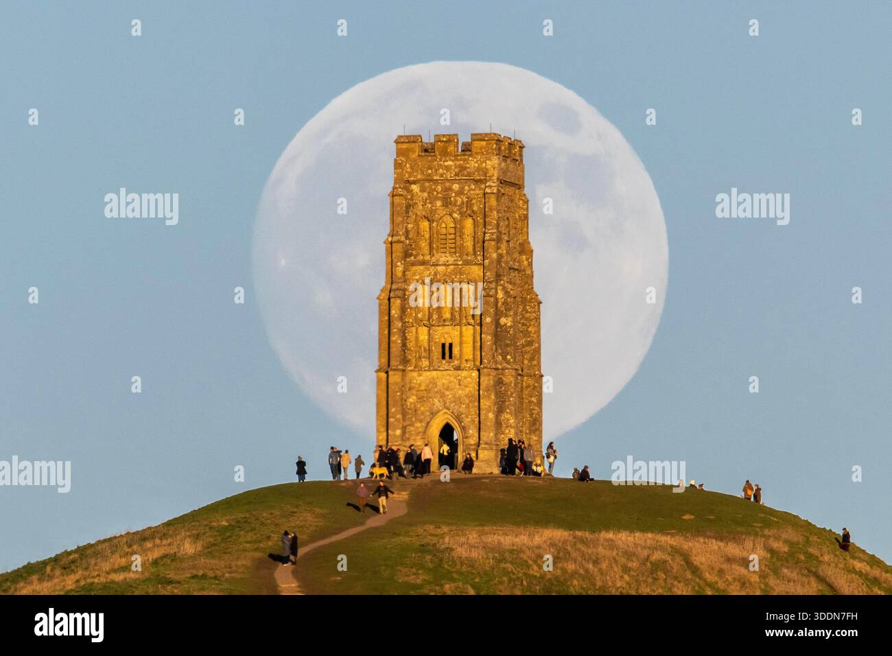 Glastonbury, Somerset, Royaume-Uni. 2 janvier 2026. Météo britannique. La lune presque pleine Wolf Moon se lève dans le ciel clair de l'après-midi de derrière la tour St Michael sur Glastonbury Tor à Glastonbury dans le Somerset comme une grande foule regarde. Ce mois la pleine lune est le 3 janvier sera une super lune. Crédit photo : Graham Hunt/Alamy Live News Banque D'Images