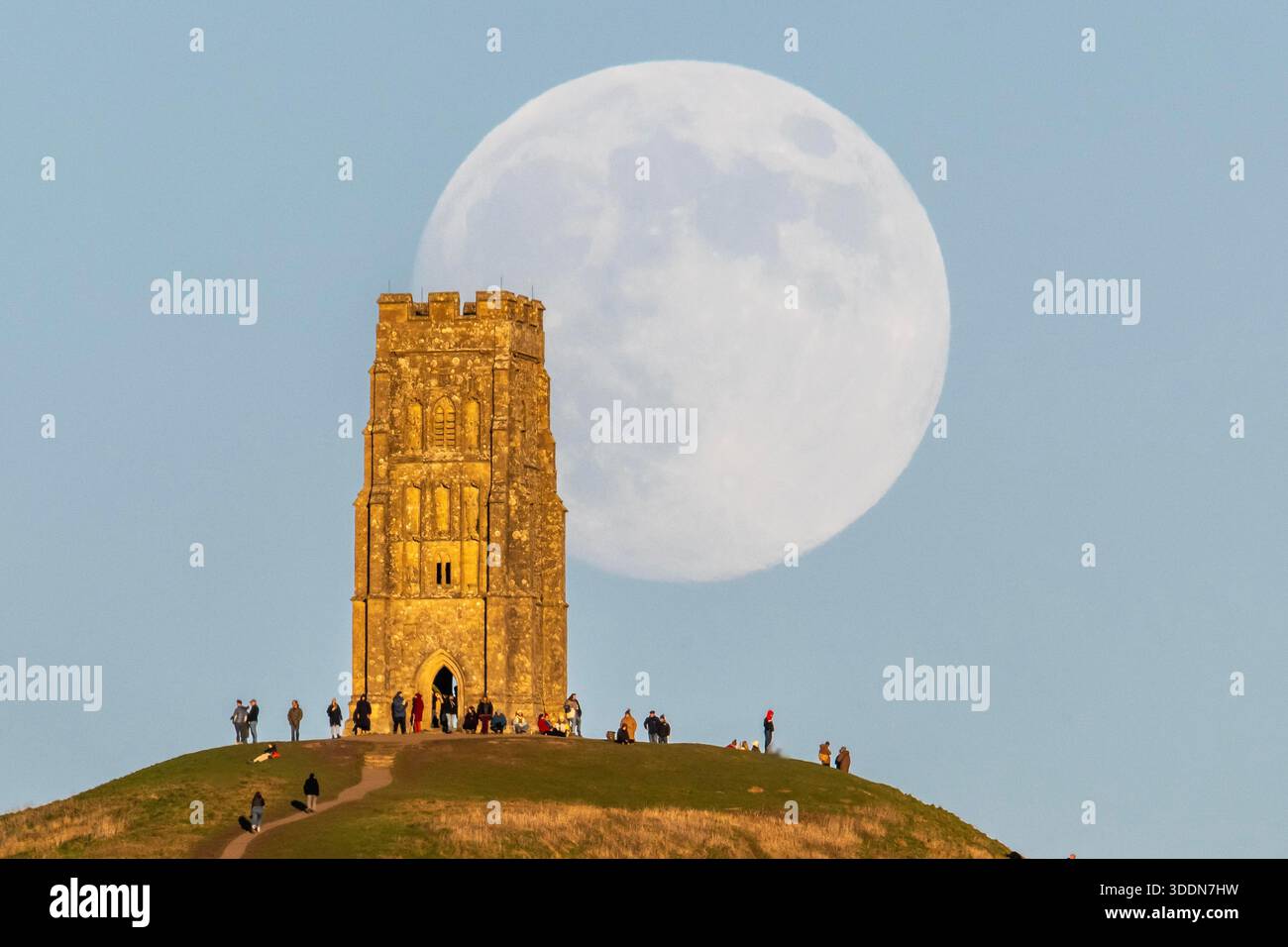 Glastonbury, Somerset, Royaume-Uni. 2 janvier 2026. Météo britannique. La lune presque pleine Wolf Moon se lève dans le ciel clair de l'après-midi de derrière la tour St Michael sur Glastonbury Tor à Glastonbury dans le Somerset comme une grande foule regarde. Ce mois la pleine lune est le 3 janvier sera une super lune. Crédit photo : Graham Hunt/Alamy Live News Banque D'Images