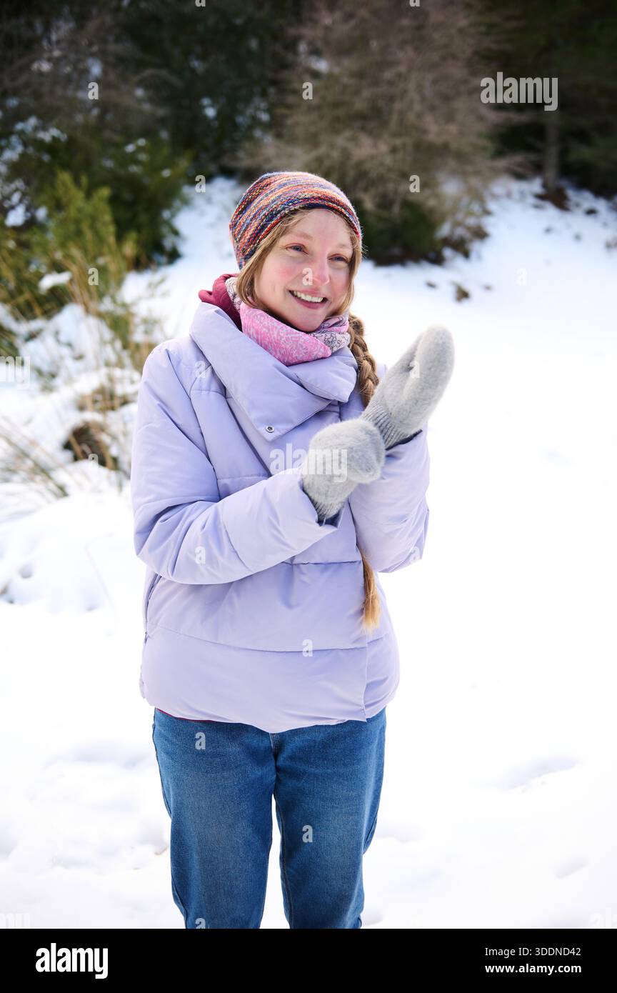 Fille souriante dans une veste d'hiver lilas, bonnet en tricot et gants se tient dans un parc enneigé, profitant du temps froid et d'un moment joyeux. Banque D'Images