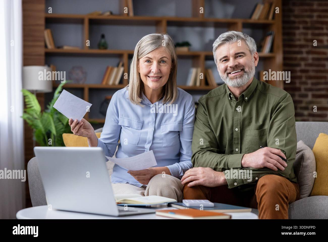 Heureux couple aîné assis sur le canapé à la maison, souriant tout en utilisant un ordinateur portable et en examinant des documents pour calculer les factures et planifier l'épargne-retraite et les finances ensemble Banque D'Images