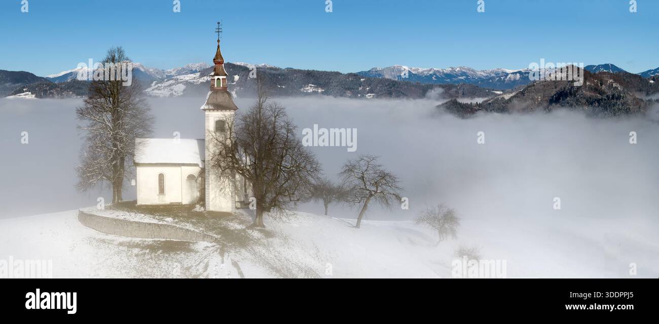 Vue panoramique de l'église Jamnik et du village dans le brouillard d'hiver : Saint Primus et l'église Felician sur une colline enneigée au lever du soleil, Alpes slovènes. Banque D'Images