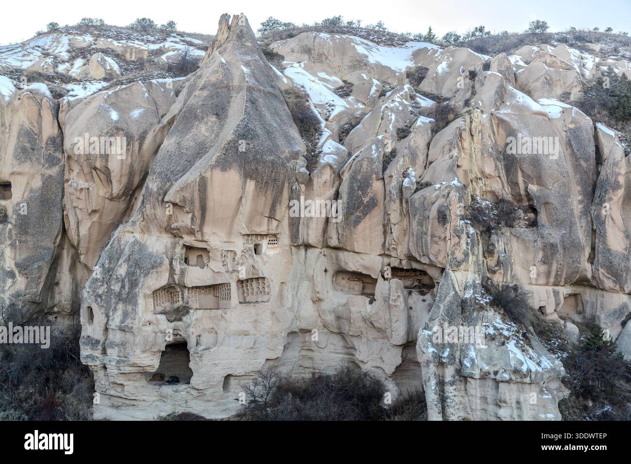 Le musée en plein air de Göreme (Müze Seyir Alanı) révèle une paroi rocheuse imposante parsemée de demeures troglodytes et de fenêtres taillées près de Göreme, en Anatolie centrale, en Turquie. Des plaques de neige persistent dans les creux du rocher de tuf crème. Le musée en plein air fait partie du site du patrimoine mondial de l'UNESCO « Parc national de Göreme et sites rupestres de la Cappadoce » depuis 1985. Banque D'Images