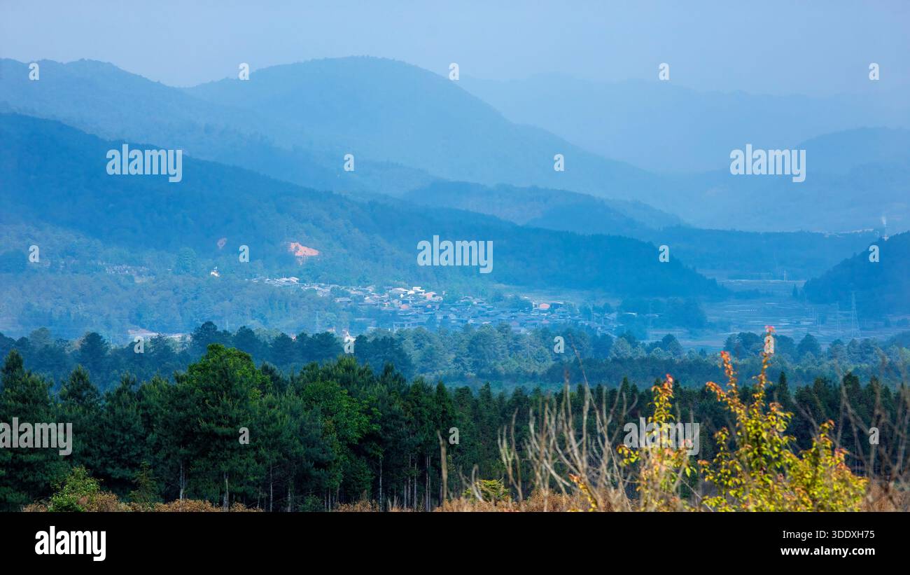 Vue lointaine d'un petit village dans une vallée de montagne entourée d'une forêt verte dense et de collines bleues en couches sous un ciel brumeux. La terre rurale tranquille Banque D'Images