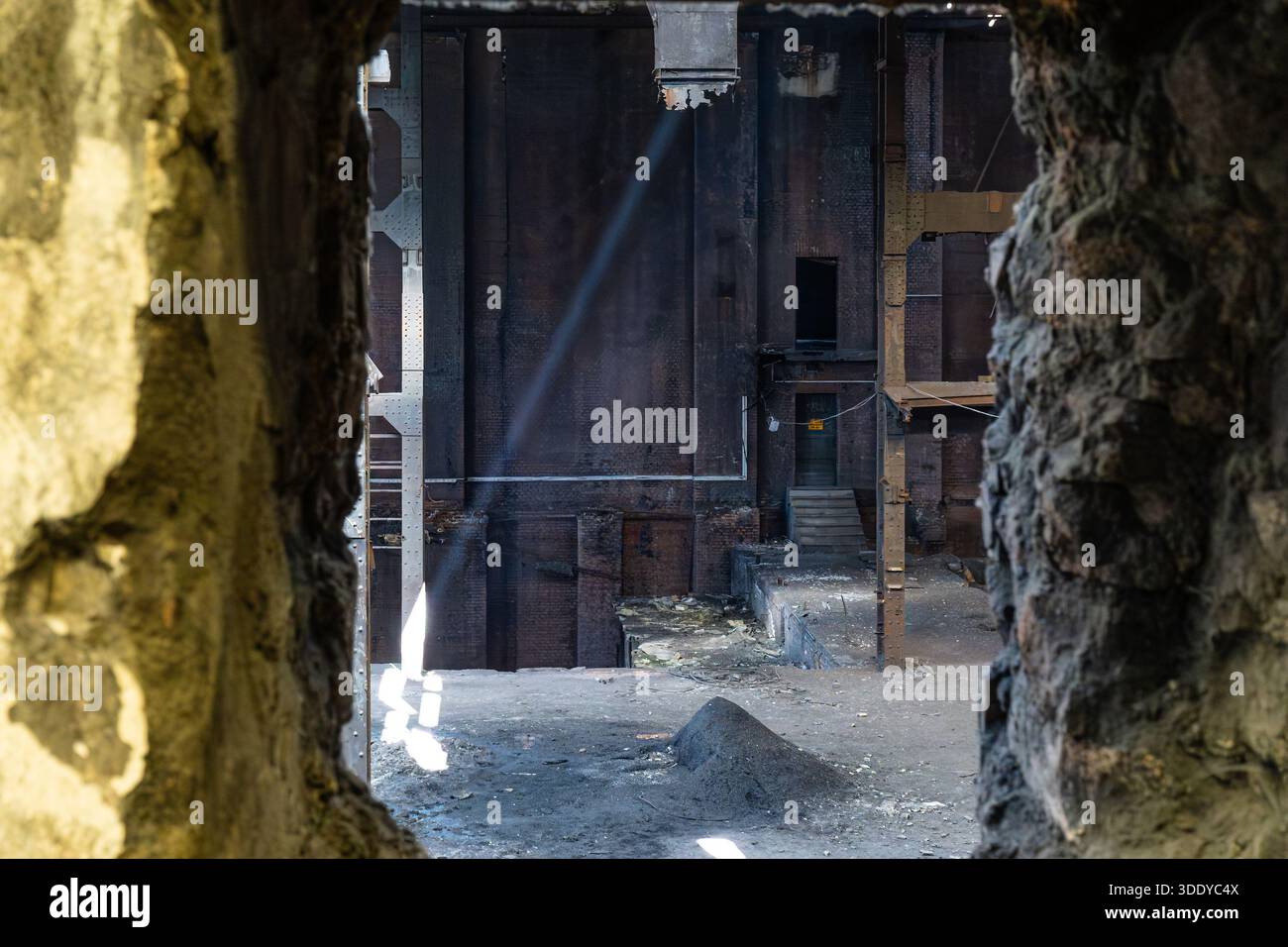 Vue intérieure d'un bâtiment industriel abandonné vu à travers un mur de béton endommagé avec des structures en acier et de la lumière naturelle Banque D'Images