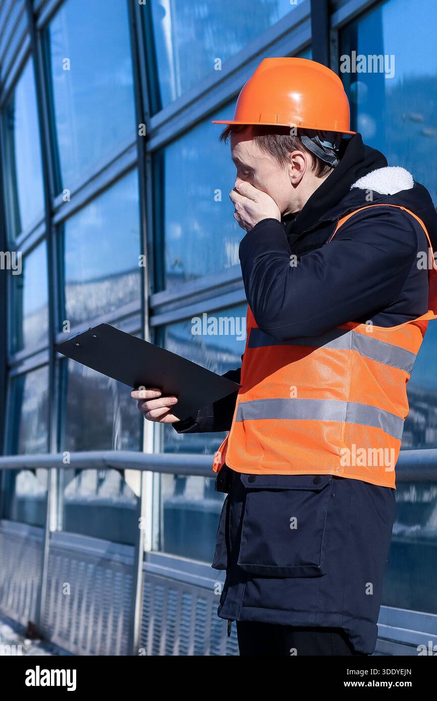 Homme de construction en tenue orange montre des notes consternées tenant près de la barrière d'hiver réfléchissante. Banque D'Images