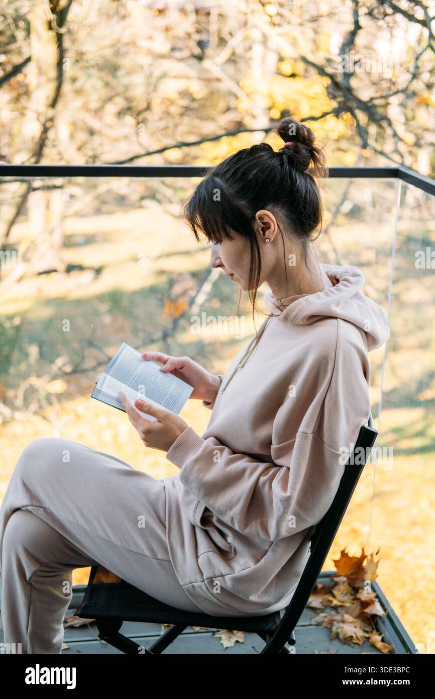 La jeune femme lit un livre en étant assise sur le balcon. Bien-être intellectuel, apprentissage tout au long de la vie, lecture attentive, vie lente. Banque D'Images