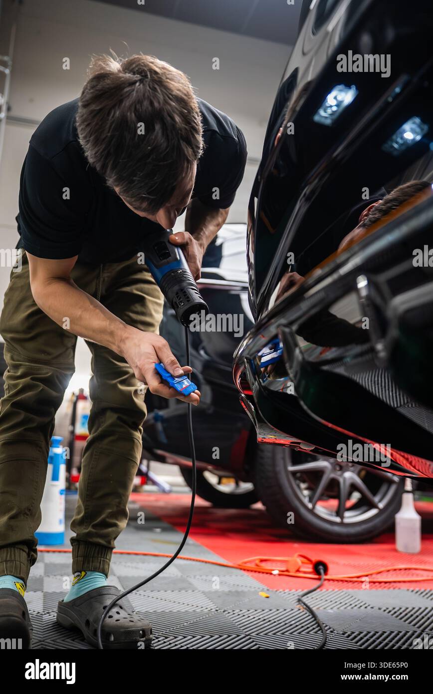 Un technicien automobile se penche sur un pare-chocs arrière noir brillant dans un atelier, à l'aide d'un pistolet thermique et d'une raclette bleue, avec des tapis de sol rouges et des outils visibles Banque D'Images