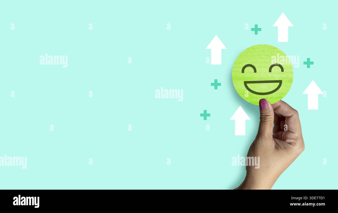 Main de femme tenant un cercle vert de papier visage souriant avec des flèches blanches et un signe plus vert autour, sur un fond vert clair. Concept de mental He Banque D'Images