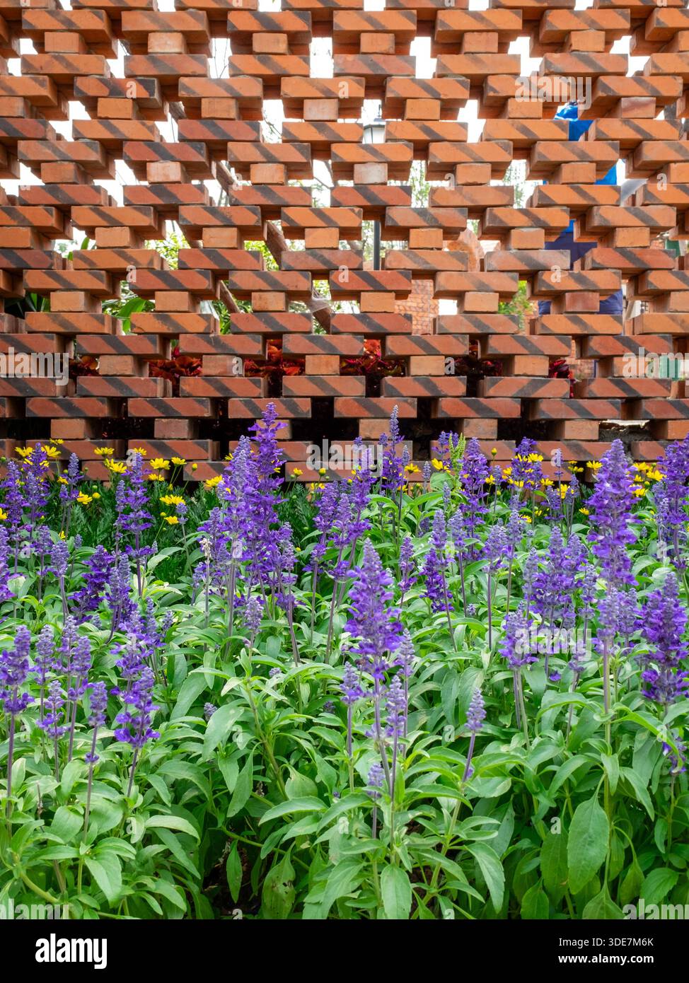 Gros plan vertical d'un mur de briques décoré de filières de vent colorées dans les espaces vides, avec des fleurs violettes en bas. L'image transmet créativ Banque D'Images