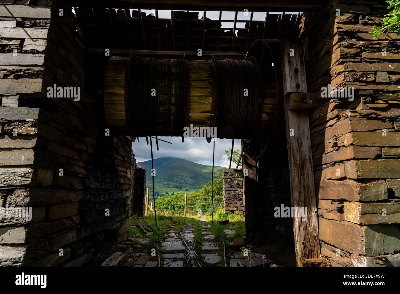 Intérieur d'un bâtiment abandonné de carrière d'ardoise avec un grand tambour d'enroulement et câble d'acier, encadrant la montagne lointaine à travers la porte Banque D'Images