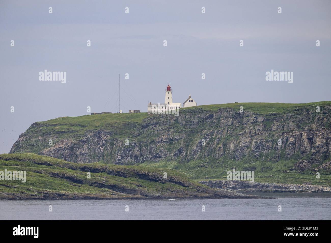 Phare blanc avec pointe rouge et antennes s'élève au-dessus des rochers abrupts sur l'île d'oiseaux de Hornoya, entouré de collines herbeuses sous un ciel clair, Vardo Banque D'Images