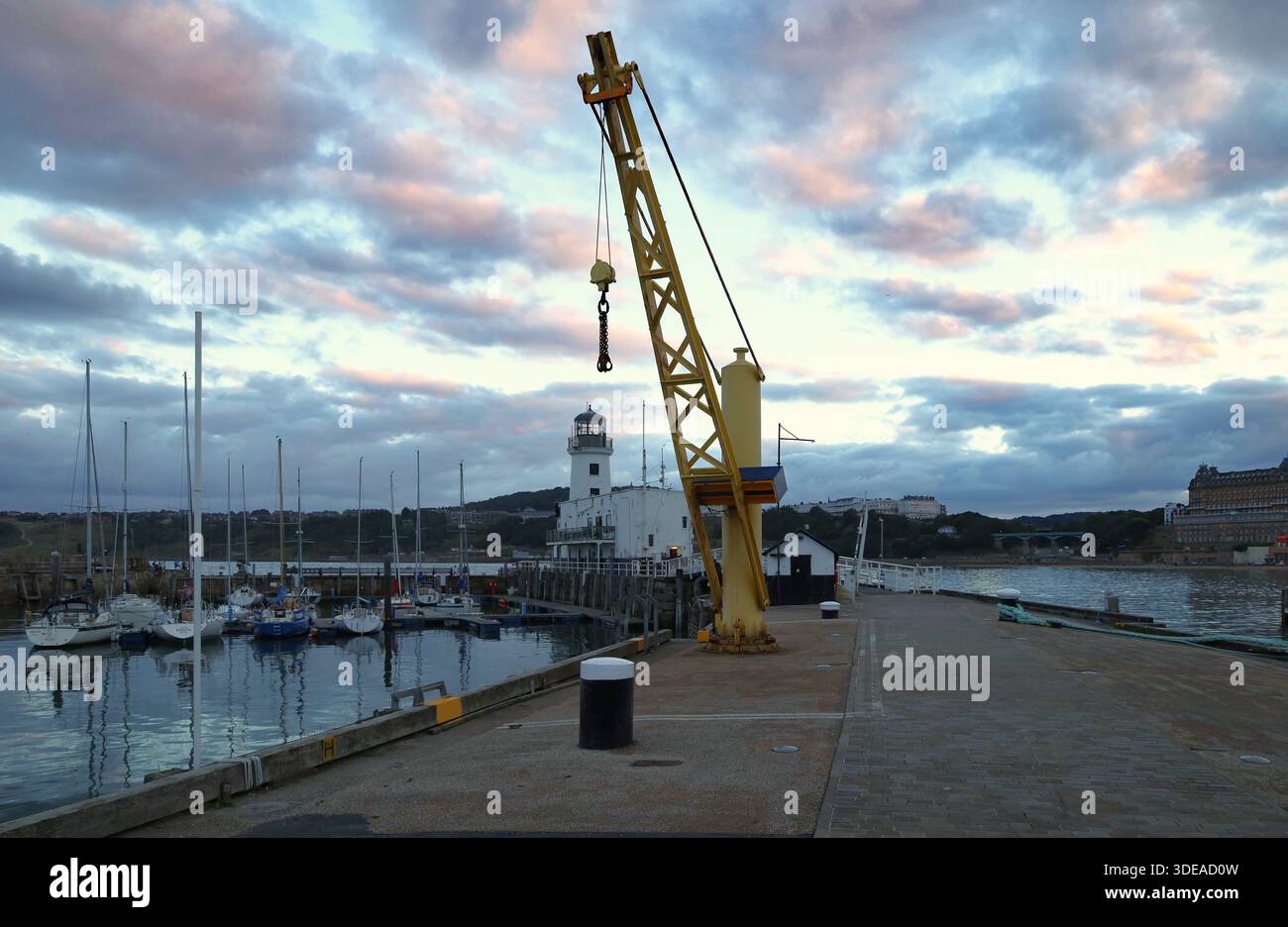 Une grue à quai sur le quai du port de Scarborough, North Yorkshire, Royaume-Uni, au coucher du soleil, avec des yachts amarrés dans la marina et un phare en arrière-plan. Banque D'Images