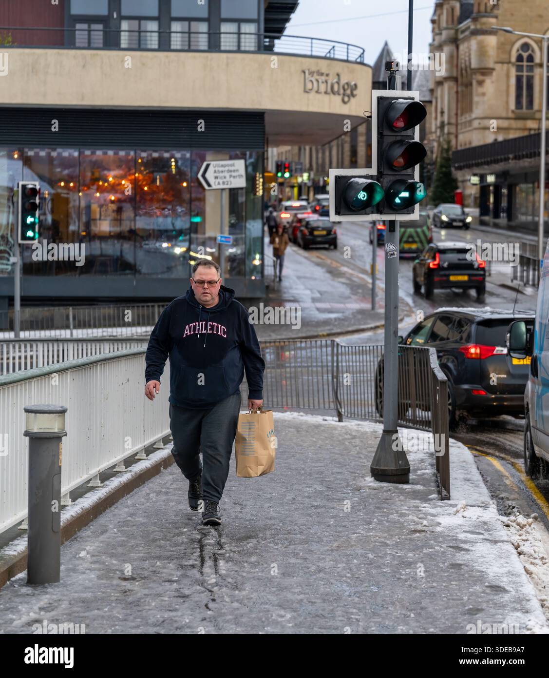Un homme marche vigoureusement sur un trottoir recouvert de neige tout en transportant un sac. Cette scène se déroule près de Ness Bridge. C'est l'hiver à Inverness. Banque D'Images