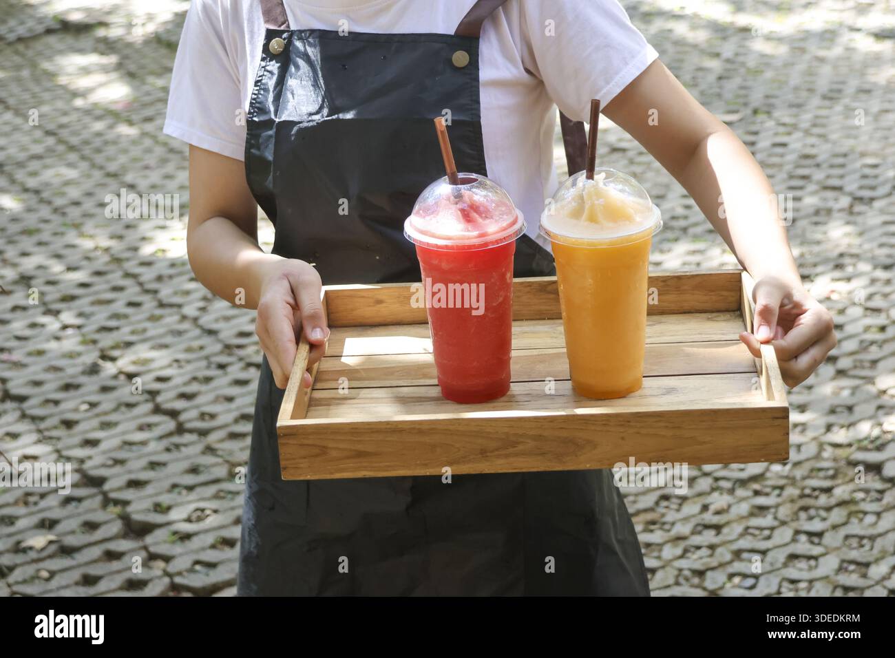 Plan recadré de serveuse portant un plateau en bois avec smoothie aux fruits rafraîchissants froids dans une tasse en plastique servant le client avec une émotion accueillante à l'extérieur Banque D'Images